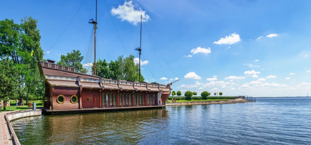 Wooden pirate-style ship moored along a grassy lakeside under a blue sky with trees nearby.