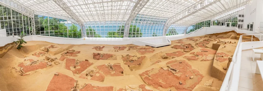 Panoramic view of an indoor archaeological excavation site inside a glass-walled conservatory with sandy trenches and scattered stones, overlooking a lake/ocean beyond the windows.