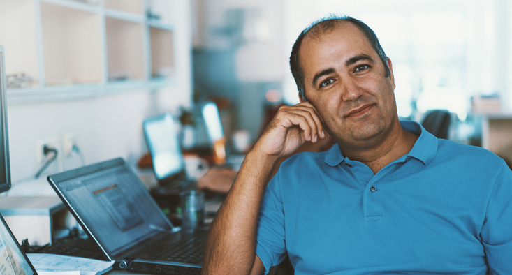 Man in a blue polo sits at a desk in an open-office, smiling at the camera with a laptop in front of him.