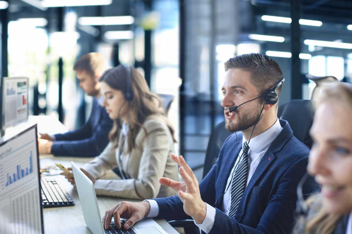 Support agents with headsets working at computers in a bright open office environment, foreground agent gesturing while on a call
