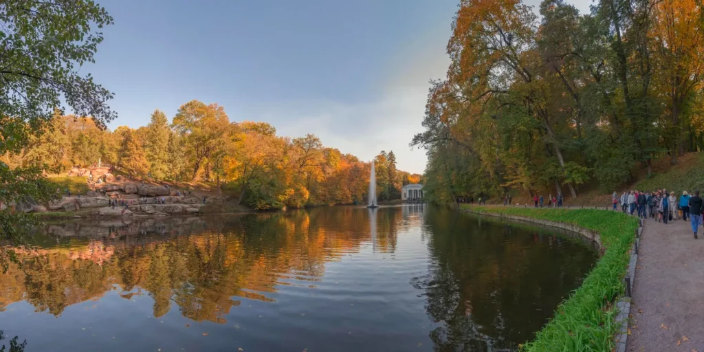 Panoramic view of a park lake in autumn with a fountain, a rocky shore on the left, and people walking a path along the right edge.