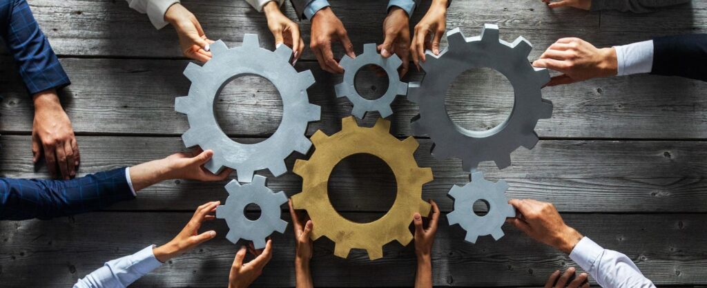Diverse hands working together to fit gray and gold gears on a wooden table.