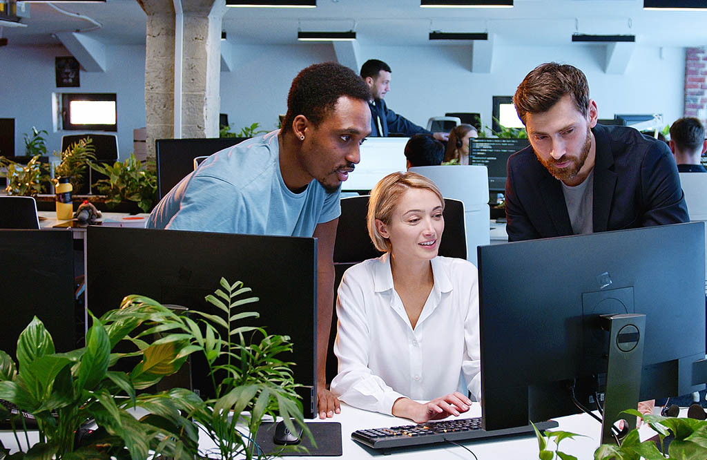 Three coworkers collaborate at a desk, looking at a computer screen amid green plants in a bright office environment.