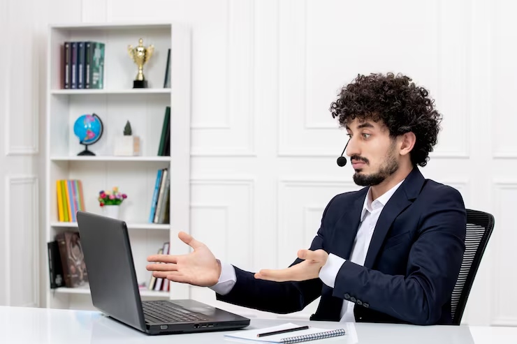 Male customer service agent in a navy suit with a headset sits at a white desk, gesturing while speaking to a client on a laptop.