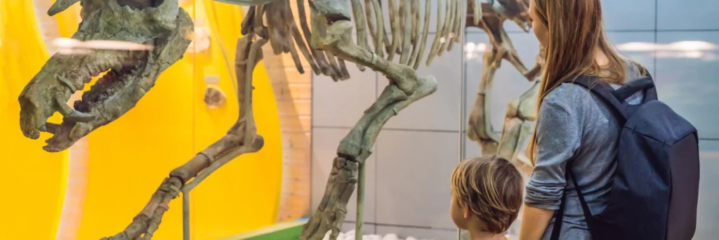 Woman with a backpack and a child observe a large dinosaur skeleton in a museum exhibit against a bright yellow background.