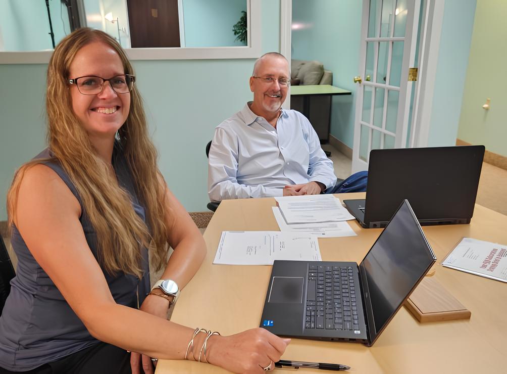 Two coworkers sit at a conference table with laptops and documents, smiling at the camera in a bright office setting.