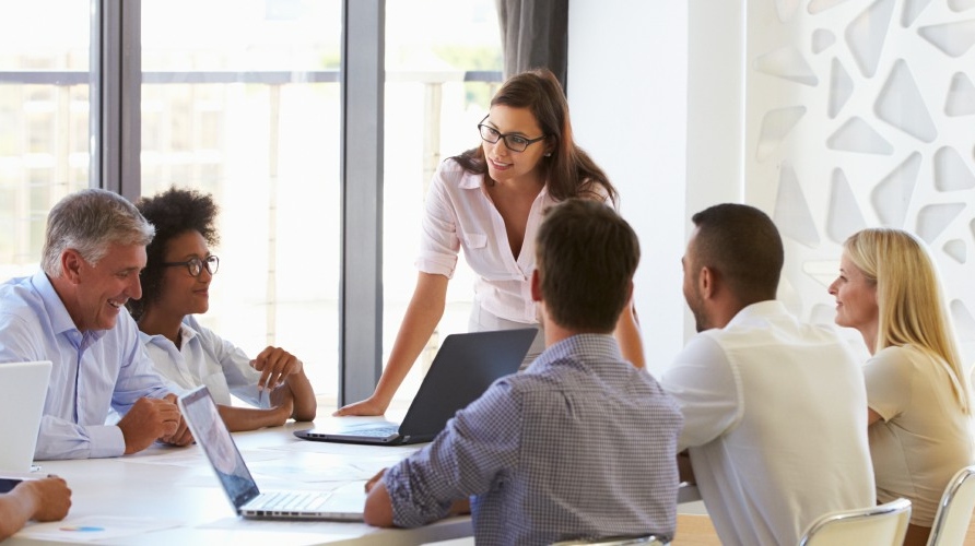 A woman stands and leans over a conference table, presenting to a diverse group of colleagues with laptops open in a bright meeting room.