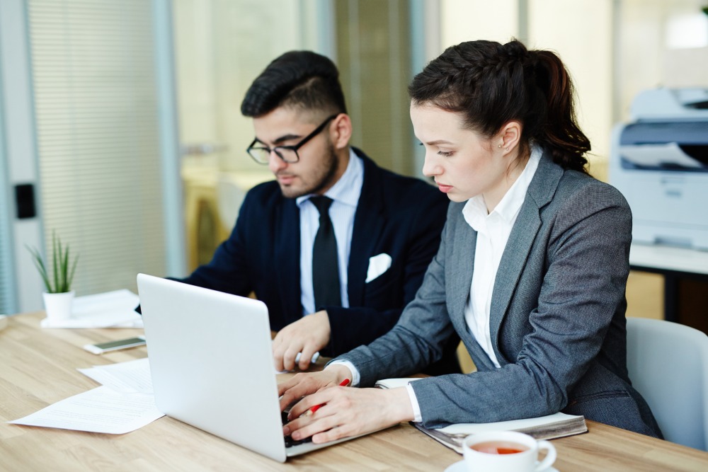 Two professionals in business attire work on a laptop at a bright office desk with papers nearby.