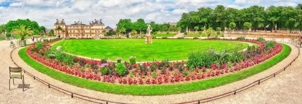 Panoramic view of a formal circular garden with a statue at the center, vibrant flower borders, and a historic building in the background.