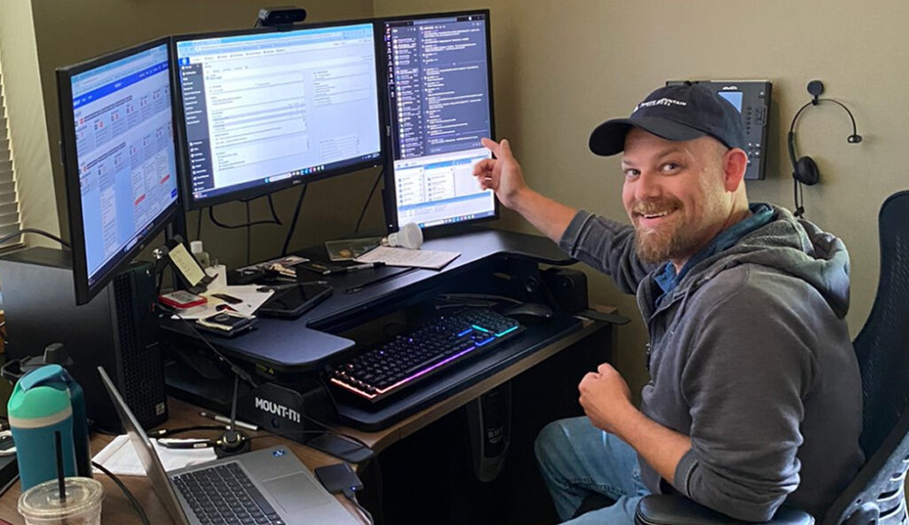 Smiling man wearing a cap sits at a desk with a three-monitor computer setup and a glowing keyboard nearby.