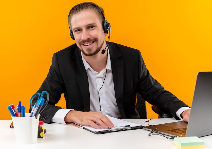 Smiling man in a suit with a headset, seated at a desk with notebook and laptop against an orange background.