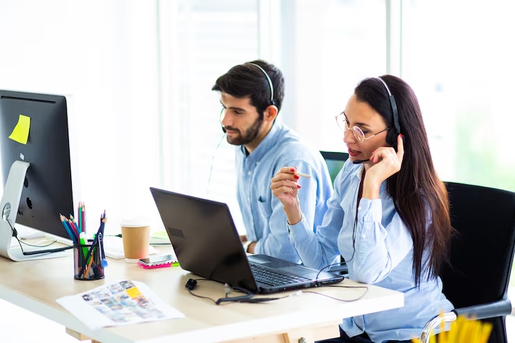 Two customer service agents with headsets at a shared desk, working on computers and a laptop.