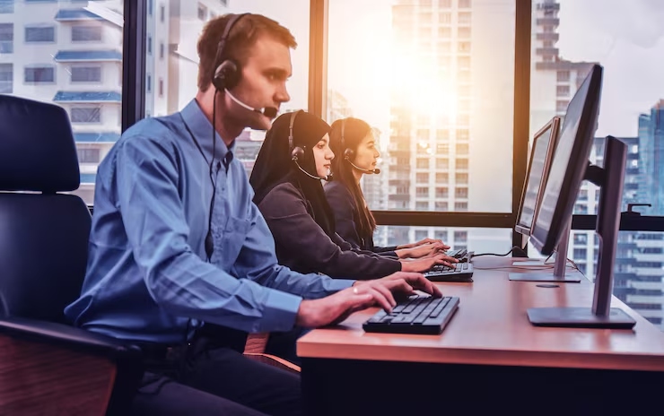 Three call center agents wearing headsets sit at desks with computers in a bright office, assisting customers.