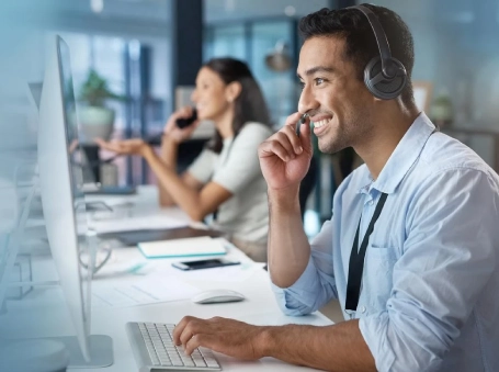 Customer service representative wearing a headset and typing at a computer in an office, with a colleague on a call in the background