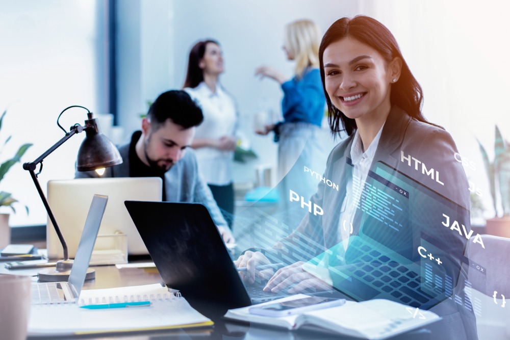 Smiling female developer at a laptop with code language overlays and colleagues working in a bright modern office behind her.