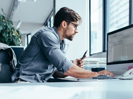 Man wearing a blue shirt sits at a desk in a bright office, using a computer and holding a smartphone.