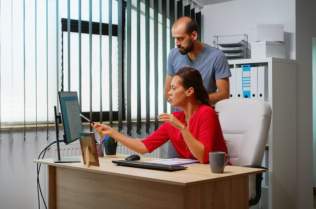 Woman in red blouse sits at a desk and points at a computer monitor while a man stands behind her, assisting in a bright office with vertical blinds.