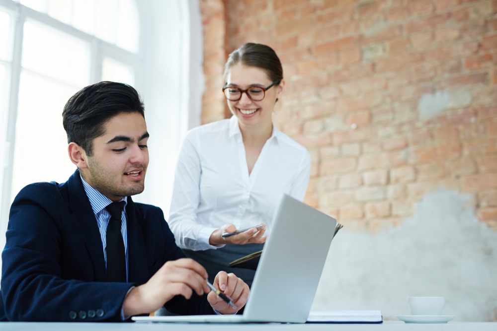 Two professionals discuss work at a laptop; man in suit writes while a woman coworker stands beside him, smiling with a notebook in hand.