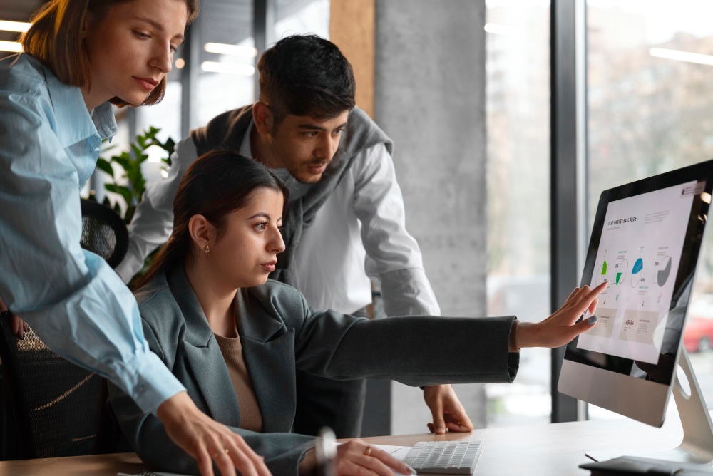 Three colleagues collaborate around a desktop monitor, with the woman in a blazer pointing at charts on a large screen.
