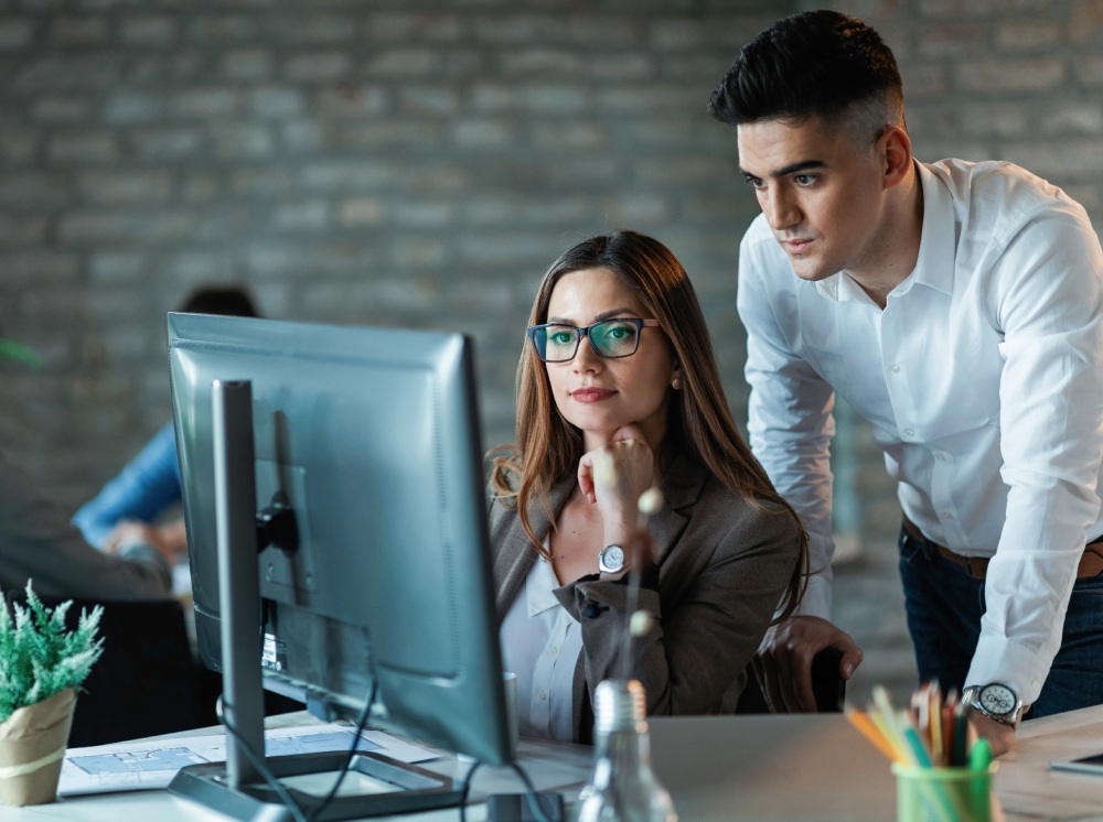 Two colleagues work together at a desk, focusing on a computer monitor in a modern office.