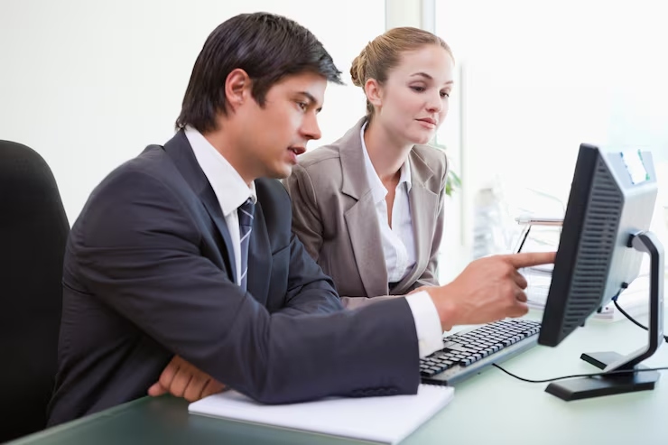 Two business colleagues collaborate at a desk, the man pointing at a computer screen as the woman looks on at a bright office.