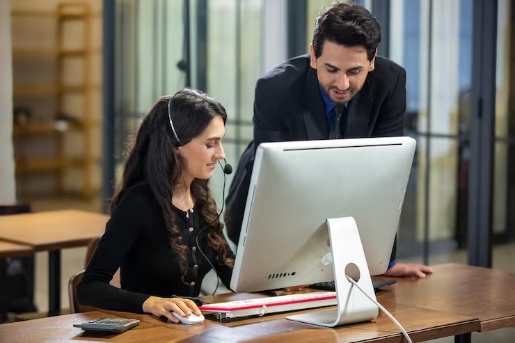 Woman wearing a headset sits at a desk using a desktop computer as a man leans in to help nearby in a modern office.