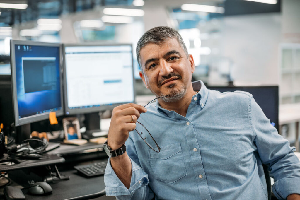 Smiling man in a light blue shirt sits at a desk with two computer monitors behind him, holding his eyeglasses near his mouth.